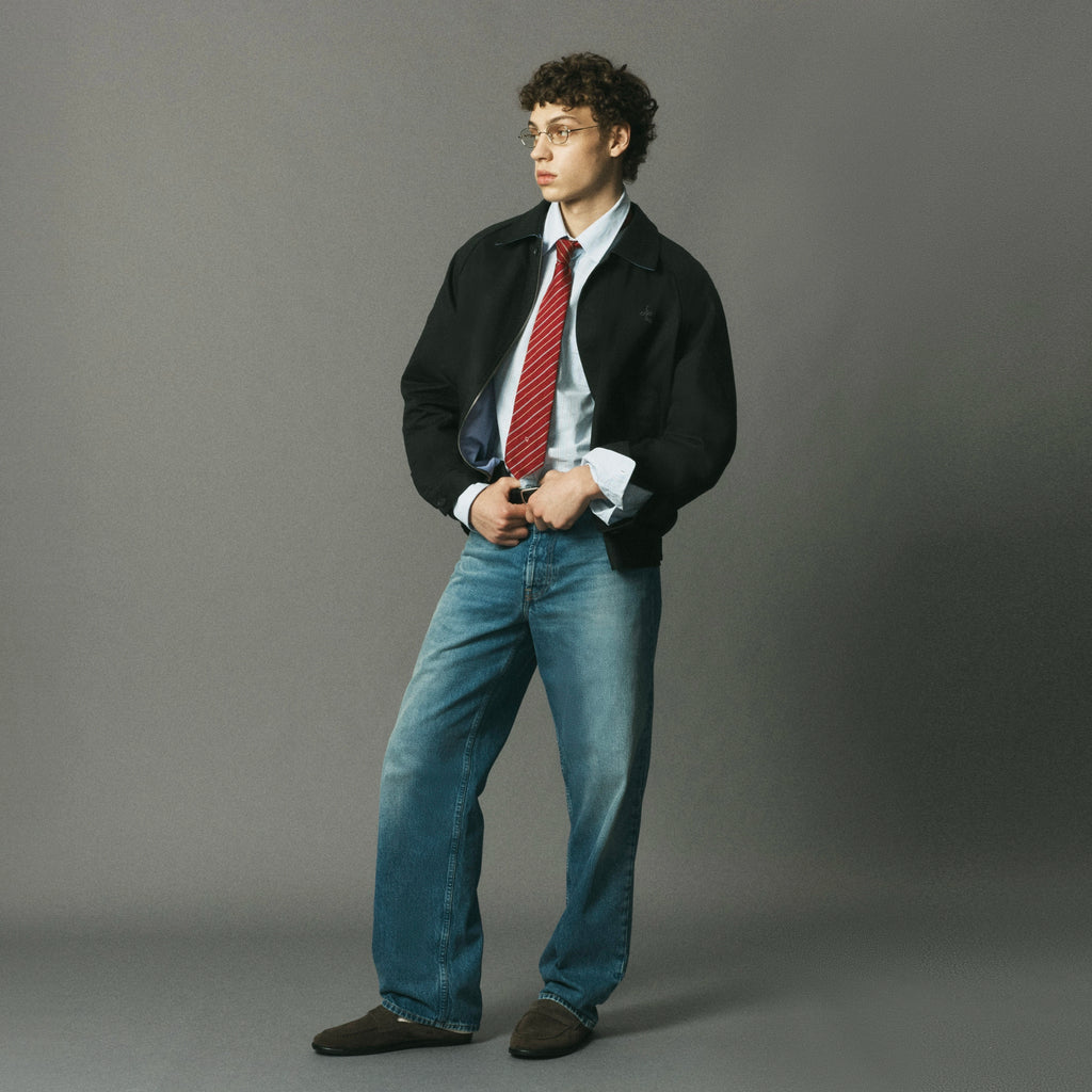 A young man wearing a black blazer, light blue dress shirt, red striped tie, blue jeans, and brown shoes, standing against a solid gray studio background.