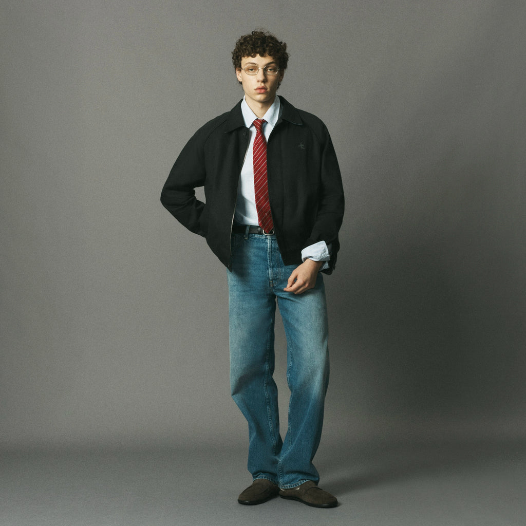 A model stands against a solid gray studio background wearing a black jacket over a white collared shirt with a red patterned tie, blue jeans, and brown shoes.