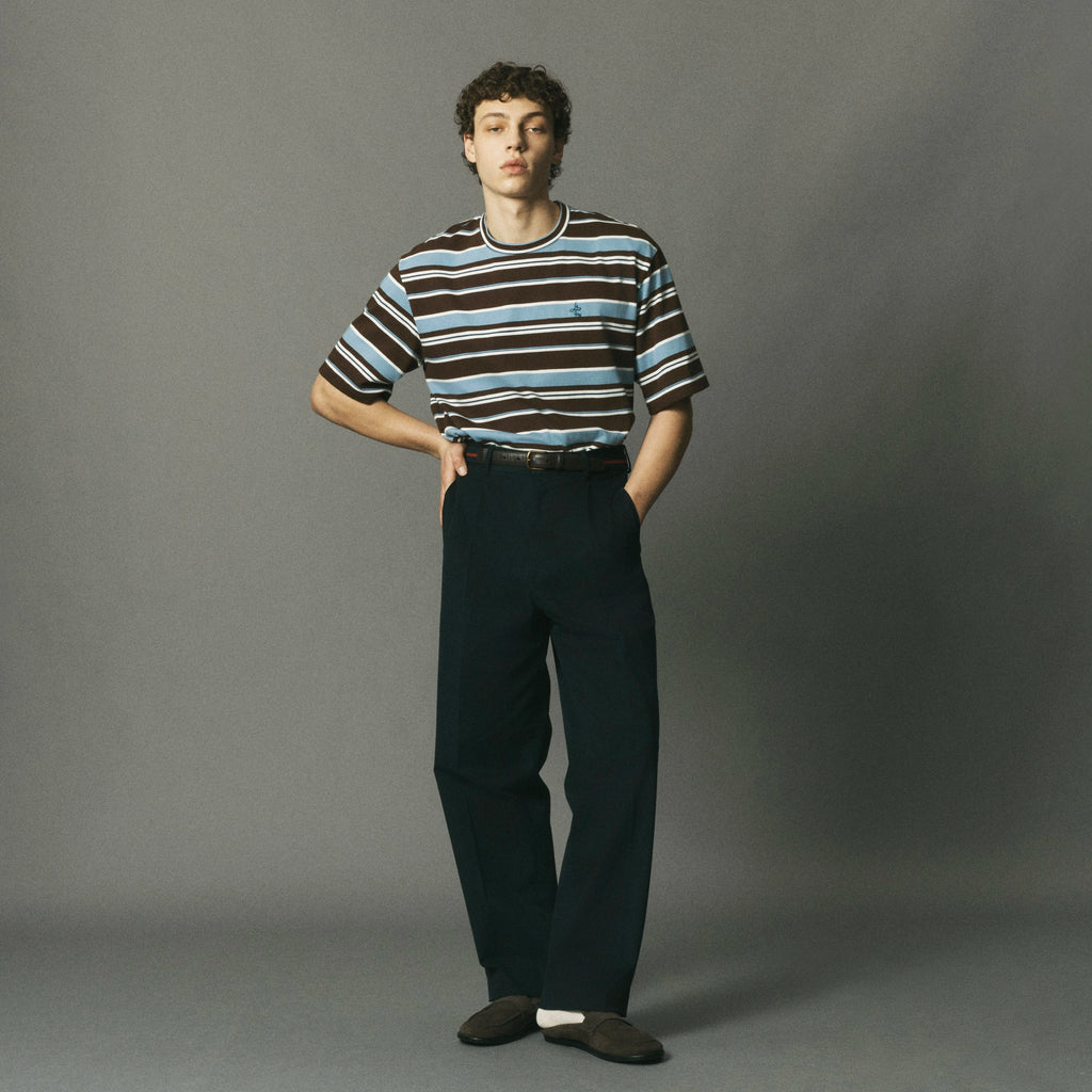 A model stands against a gray studio background wearing a short-sleeve crewneck t-shirt with brown, light blue, and white horizontal stripes, paired with dark teal trousers and dark brown knit slip-on shoes.