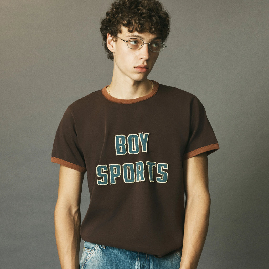 A young man with curly hair and glasses wearing a dark brown ringer t-shirt with 'BOY SPORTS' printed on the front in teal and white, paired with blue jeans, against a solid gray studio background.