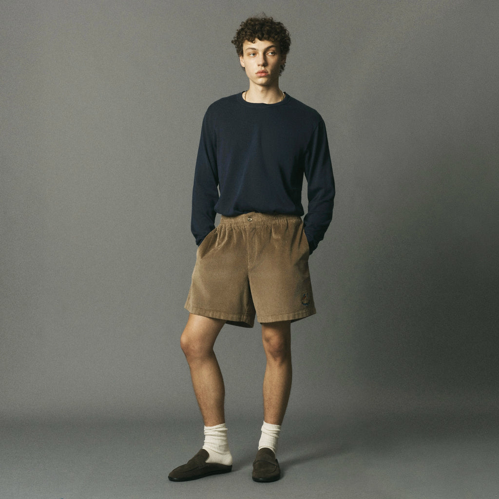 A young man stands against a plain gray studio background wearing a dark navy long-sleeve crewneck sweater, tan corduroy shorts, white socks, and dark loafers.