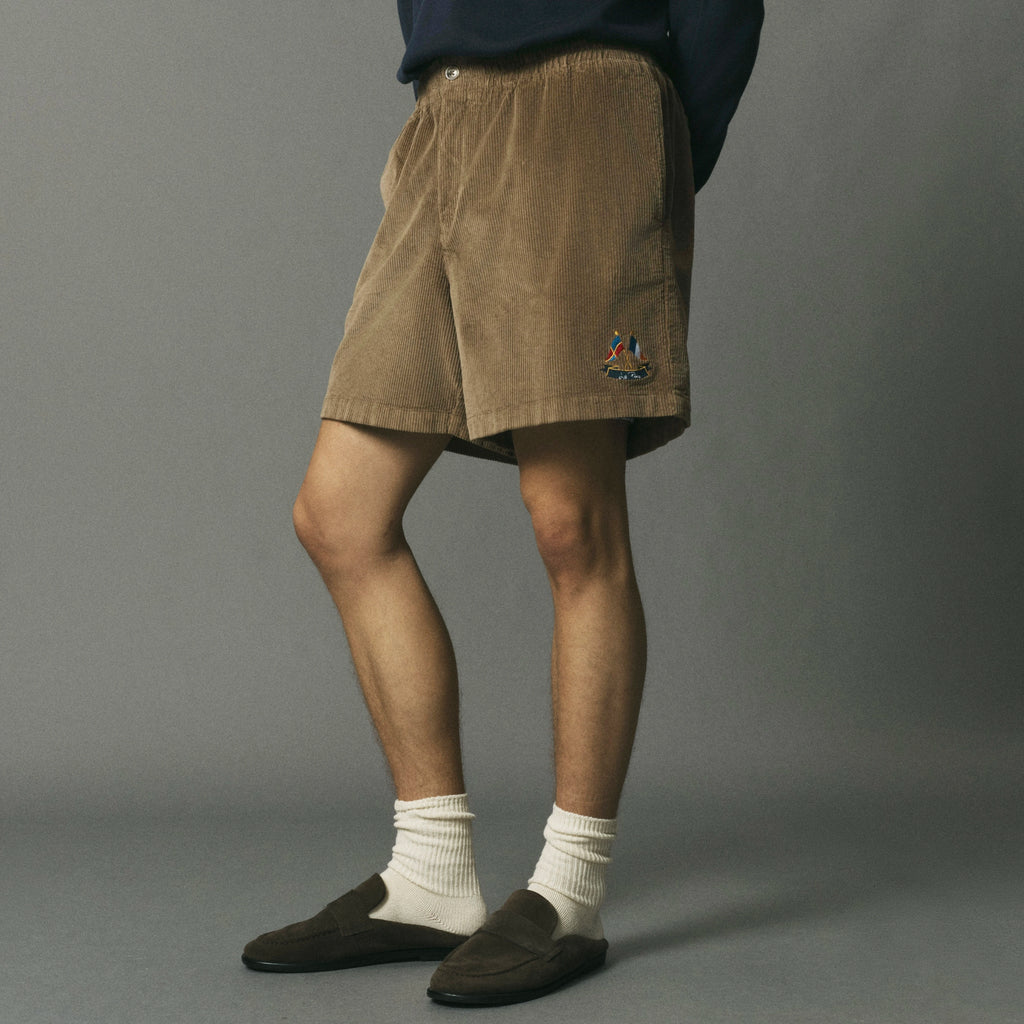 A person wearing tan corduroy shorts with a small embroidered logo on the lower left leg, styled with white crew socks and dark brown slip-on loafers against a plain gray studio background.