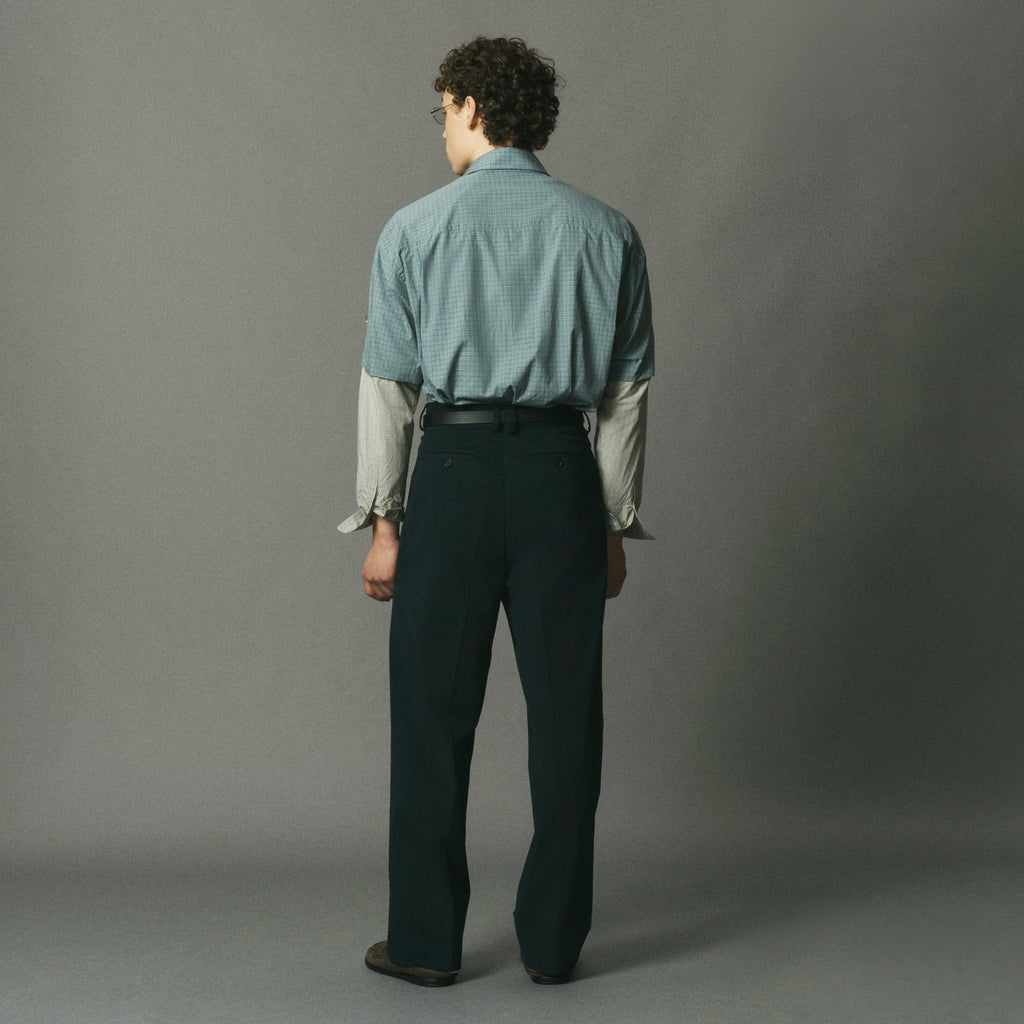 A man stands with his back to the camera wearing a light blue and white micro-check patterned button-down shirt tucked into dark navy trousers, set against a plain gray studio background.