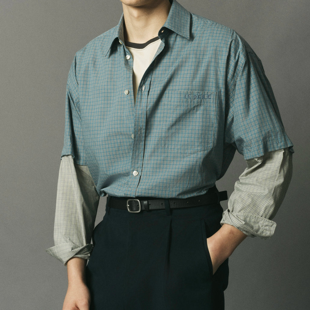 A man wearing a blue-green and white checkered button-down shirt with long sleeves rolled up, layered over a white undershirt and paired with dark trousers and a black belt, against a solid gray background.