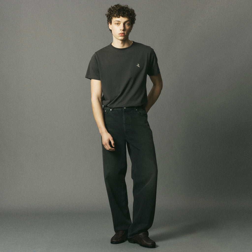A model stands against a solid gray studio background wearing a dark charcoal gray crewneck t-shirt with a small embroidered bird logo on the left chest, paired with dark charcoal gray jeans and dark brown leather shoes.