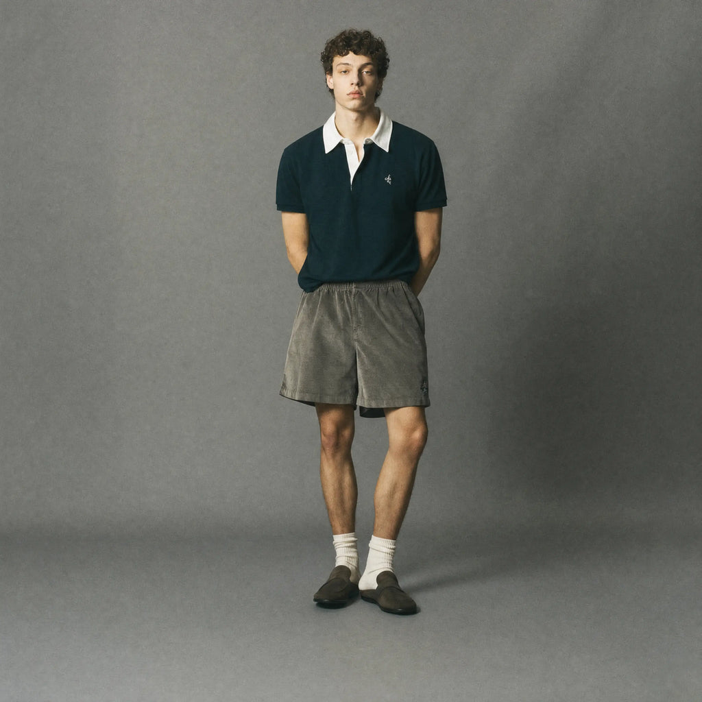 A man stands against a plain gray studio background wearing a dark navy polo shirt with a white collar, gray shorts, white socks, and dark loafers.