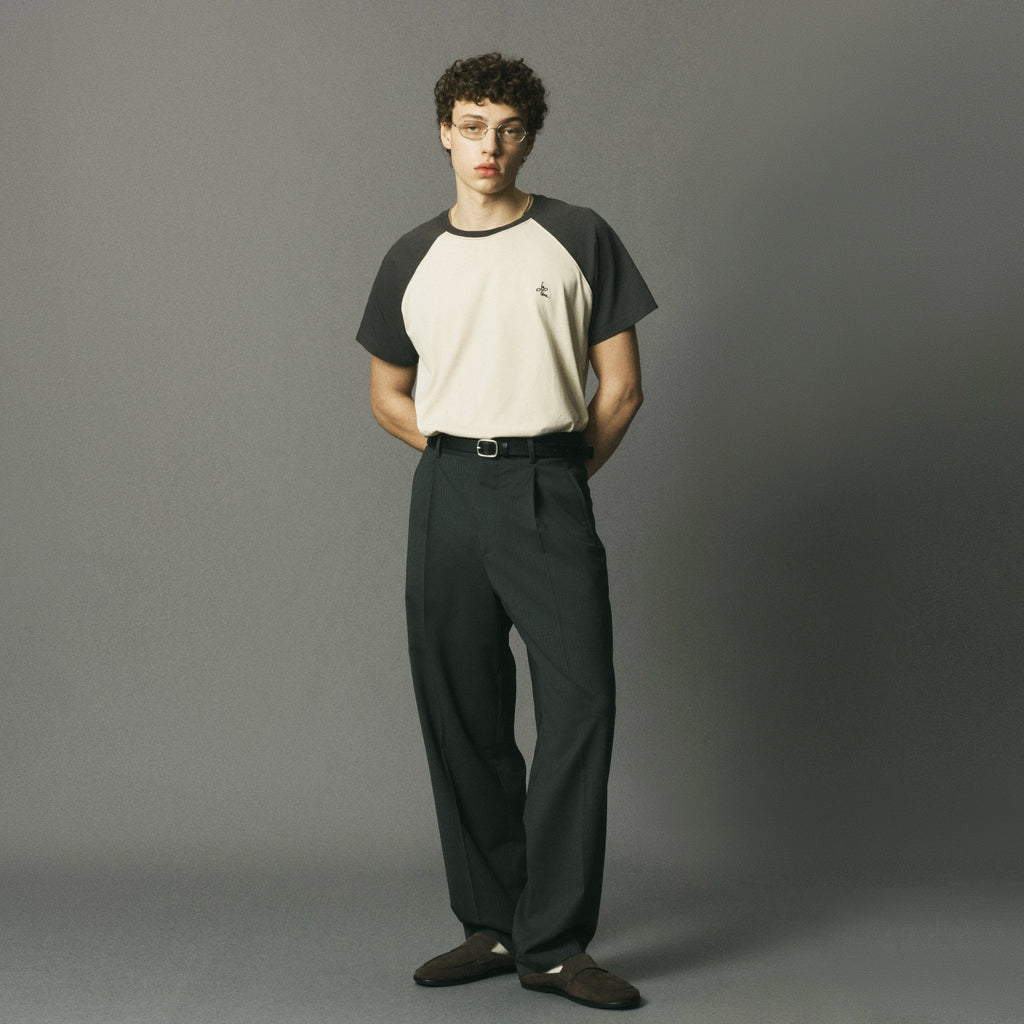 A man wearing a short-sleeve raglan t-shirt with a cream body and dark gray sleeves, paired with dark gray trousers and brown shoes, standing against a solid gray studio background.