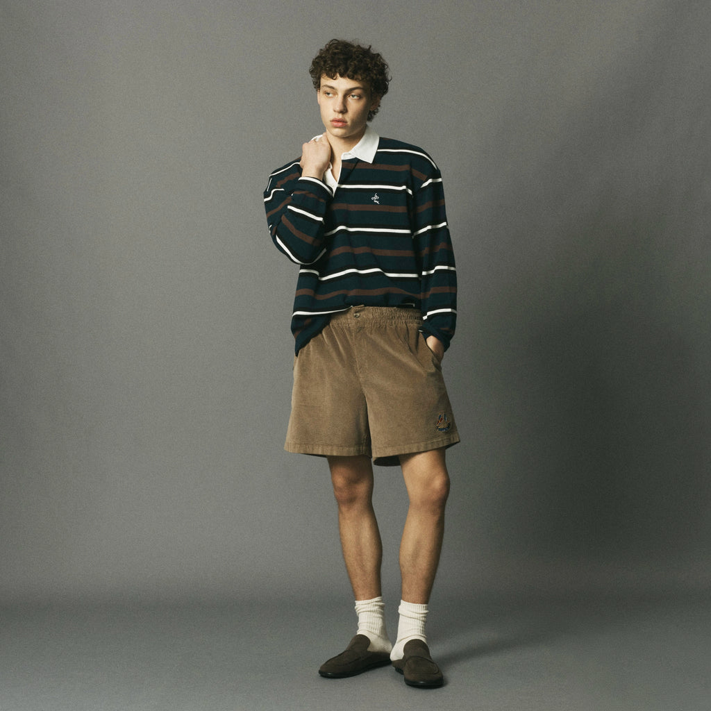 A man stands against a plain gray studio background wearing a dark navy and navy striped long-sleeve rugby shirt with a white collar, brown corduroy shorts, white socks, and dark loafers.