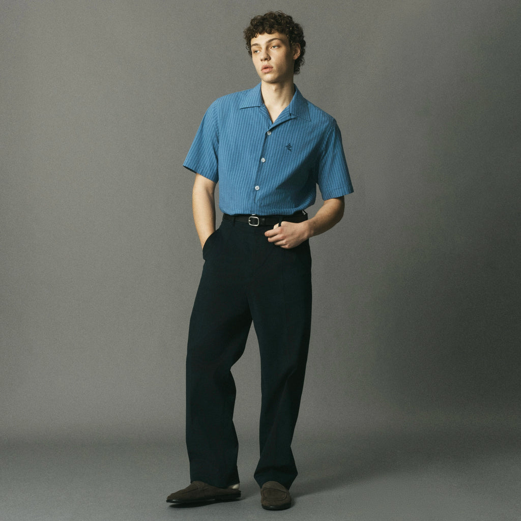 A model stands against a gray studio background wearing a short-sleeve blue button-up shirt with a subtle vertical stripe pattern, paired with dark trousers and brown shoes.