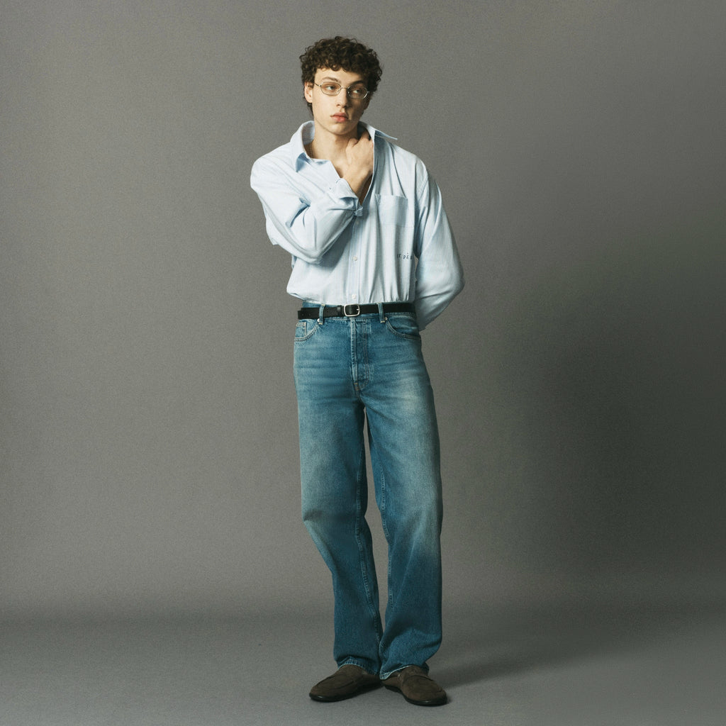 A man wearing a light blue button-down shirt, blue jeans, and dark brown shoes stands against a solid gray studio background.