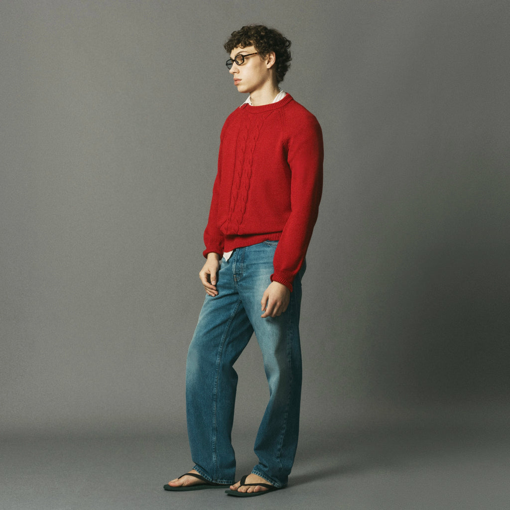 A man wearing a red cable-knit sweater, blue jeans, and black sandals stands in profile against a solid gray studio background.