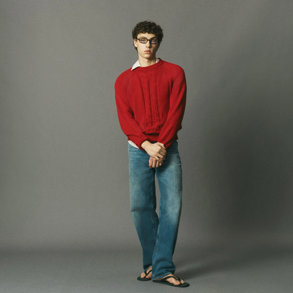 A man wearing a red cable-knit sweater, blue jeans, and black sandals stands against a solid gray studio background.
