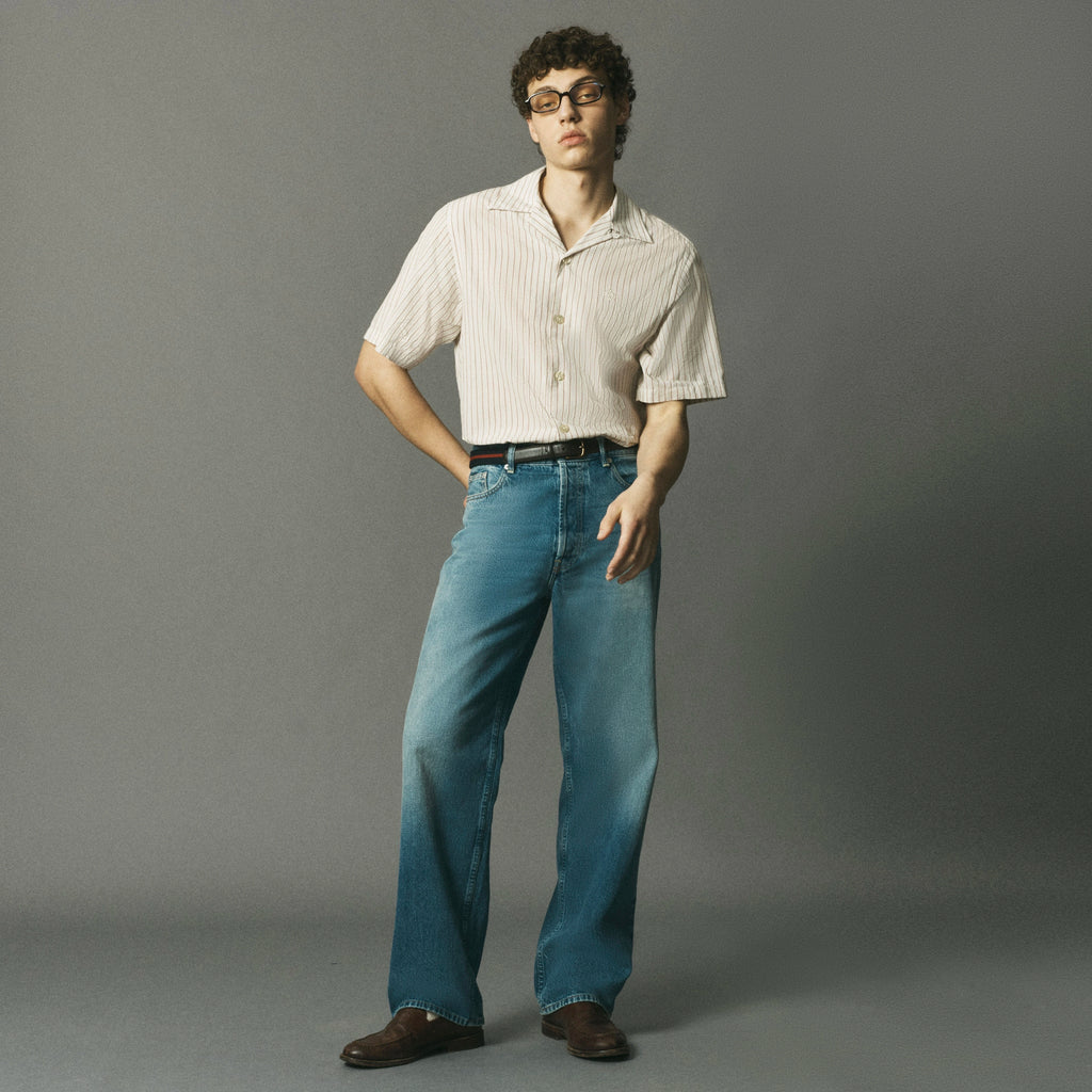 A model wearing a cream and white vertically striped short-sleeve button-up shirt, blue wide-leg jeans, a black belt, and brown leather shoes, standing against a solid gray studio background.