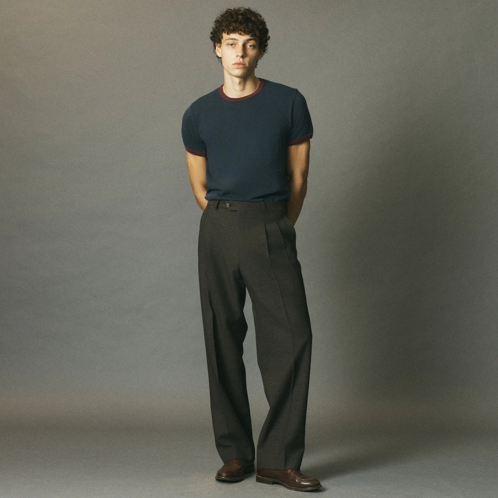A man stands against a plain gray studio background wearing a dark navy short-sleeve crewneck t-shirt, dark charcoal gray pleated trousers, and brown leather shoes.