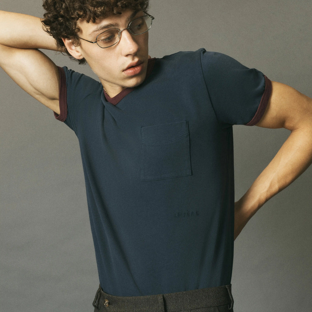 A man wearing a dark navy blue ringer t-shirt with burgundy trim on the collar and sleeve cuffs, posed against a solid gray studio background.