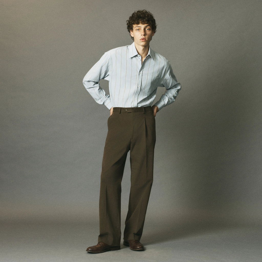 A man stands against a plain gray studio background wearing a light blue and white vertically striped button-down shirt, dark brown pleated trousers, and brown leather dress shoes.