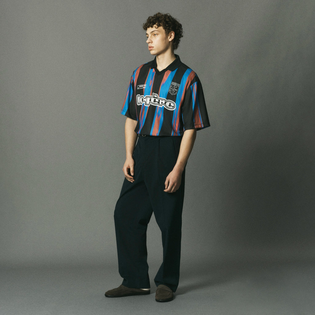 A model stands in a studio wearing a cropped short-sleeve soccer jersey with vertical navy, red, and orange stripes, paired with dark wide-leg trousers and brown shoes. The jersey features the word 'Cruze' across the chest.