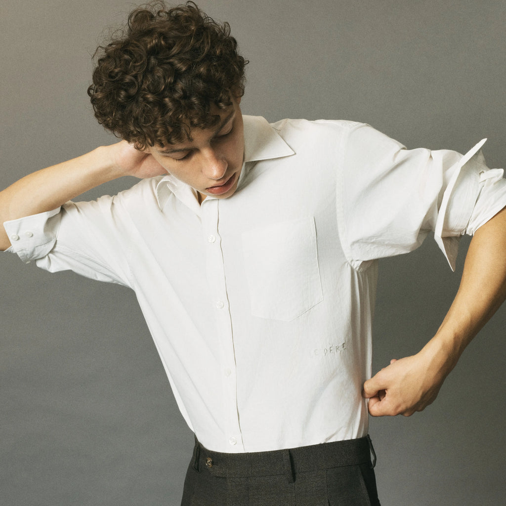 A man with curly brown hair wearing a white short-sleeve button-up shirt and dark trousers, posing against a solid gray studio background.